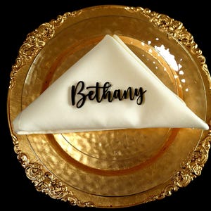 Puede incluir: Un plato dorado con un borde adornado sostiene una servilleta blanca doblada. El nombre "Bethany" está escrito en escritura negra sobre la servilleta. Un tenedor y un cuchillo dorados se colocan a la derecha.