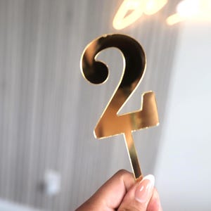 以下が含まれることがあります： 金色の数字24のケーキトッパー。数字は光沢のある反射素材で作られています。トッパーは人が持っています。背景はぼやけており、明るいグレーと白です。