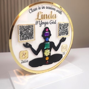 May include: A round, white and gold sign with the text "Class is in session with Linda #Yoga Girl". The sign features a black silhouette of a person in a yoga pose, with colorful gems representing chakras. Two QR codes and social media icons are also included.