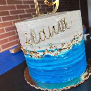 May include: A two-tiered cake with white and blue frosting. The top tier is white with the name "Alana" in gold script. The bottom tier is blue with a wave-like design. A gold "19" cake topper is on top.
