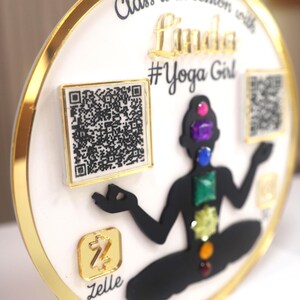 Könnte beinhalten: Ein weiß-goldenes kreisförmiges Dekorationsobjekt mit dem Text "Class is in session with Linda #Yoga Girl". Es zeigt eine schwarze Silhouette einer Person in einer Yoga-Pose, mit farbigen Edelsteinen, die Chakren darstellen, und zwei QR-Codes.