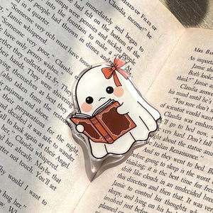 May include: A clear acrylic ghost-shaped pin, reading a book with a brown cover. The ghost has a small orange bow. The pin is on top of an open book. The ghost has a cute expression.