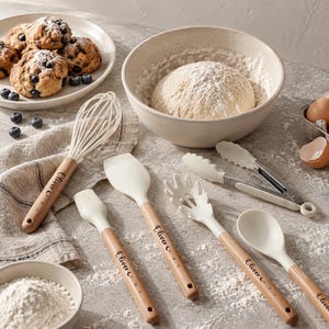May include: A set of kitchen utensils including a whisk, a basting brush, a spatula, a pasta server, a spoon, and tongs, all featuring light wood handles with the name Oliver C. engraved on them and white silicone heads.