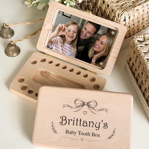 May include: A light-colored wooden baby tooth box with a hinged lid featuring a printed bow, leaf accents, and the text "Brittany's Baby Tooth Box". The interior contains circular slots for teeth and a small glass vial for storage.