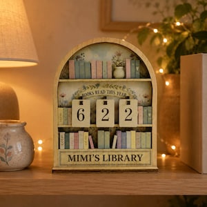 May include: An arched wooden bookshelf display featuring three rows of miniature books in pastel colors. The center row displays three hanging wooden tiles showing the number 622. The text "BOOKS READ THIS YEAR" and "MIMI'S LIBRARY" are printed on the frame.
