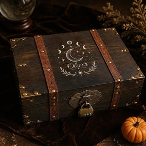 May include: A dark brown wooden chest with two vertical brown leather-like straps, brass-toned metal corner accents, and a combination padlock. The lid features a white engraved moon phase design, stars, and the name Oliver in cursive script.