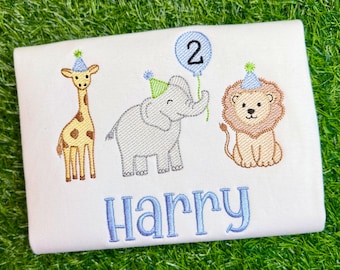 May include: A white t-shirt featuring an embroidered design of a giraffe, a gray elephant, and a lion, each wearing a small party hat. The elephant holds a blue balloon with the number 2 inside. The name Harry is embroidered in blue below.