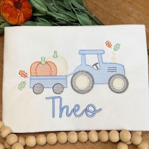 May include: A white fabric surface featuring an embroidered light blue tractor pulling a trailer filled with an orange pumpkin and a white pumpkin. The name Theo is embroidered in light blue script below the tractor. Small orange and green leaves are scattered around the design.