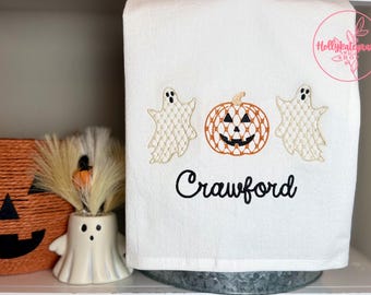 Könnte beinhalten: Ein weißes Küchentuch mit einer Stickerei von zwei Geistern, die einen zentralen orangefarbenen Kürbis mit einem schwarzen Kürbisgesicht flankieren. Der Name Crawford ist in schwarzer Schreibschrift unter dem Kürbismotiv eingestickt.