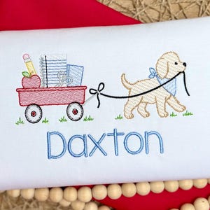 May include: A white shirt featuring an embroidered design of a light brown puppy wearing a blue bandana, pulling a red wagon filled with a red apple, a yellow pencil, and a notebook. The name Daxton is embroidered below in blue letters.