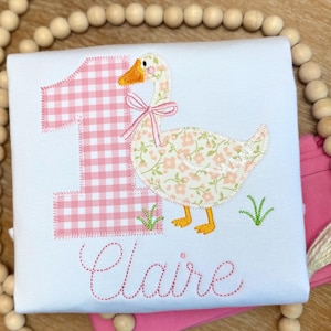 May include: White t-shirt with a pink gingham number 1 applique and a floral goose design. The goose has a pink bow and is standing on green grass. The name "Claire" is embroidered in pink script.