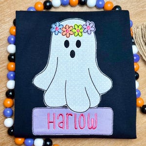 May include: Black square fabric with a white ghost applique wearing a flower crown. The ghost has a polka dot pattern. The name "Harlow" is embroidered in pink on a purple rectangle. Decorative beads surround the fabric.