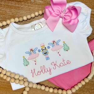 May include: White short-sleeved shirt with a Nutcracker and fairy embroidery design, pink pants, and a large pink bow. The shirt features the name "Holly Kate" in pink script. The embroidery includes a Christmas tree and snowflakes.