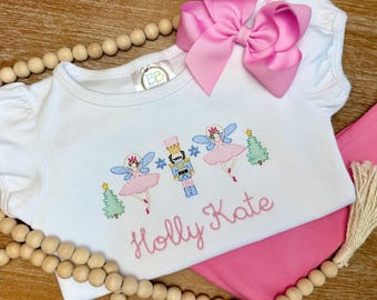 May include: White short-sleeved shirt with a Nutcracker and fairy embroidery design, pink pants, and a large pink bow. The shirt features the name "Holly Kate" in pink script. The embroidery includes a Christmas tree and snowflakes.