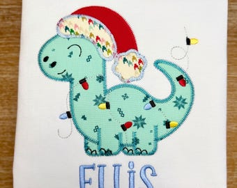 May include: A white t-shirt featuring a light blue dinosaur wearing a red Santa hat. The dinosaur is decorated with Christmas lights and the name "ELLIS" is embroidered in blue below the dinosaur.