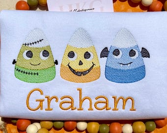 May include: A white, embroidered Halloween-themed design featuring three candy corn characters with smiling faces. The candy corn are in green, yellow, and blue. The name "Graham" is embroidered in orange below the candy corn.