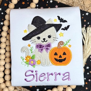 May include: A white t-shirt featuring an embroidered design of a light gray cat wearing a black witch hat and a purple bow tie. The cat is next to an orange jack-o'-lantern, yellow flowers, and two black bats. The name Sierra is embroidered in purple.