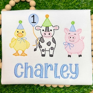 May include: A white shirt featuring an embroidered design of a yellow duck, a black and white cow, and a pink pig, all wearing party hats. A blue balloon with the number 1 is above the cow. The name Charley is embroidered in blue below.