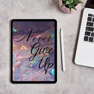 Może przedstawiać: Tablet cyfrowy wyświetla słowa "Never Give Up" w eleganckim skrypcie na holograficznym tle. Biały rysik spoczywa obok tabletu, z laptopem i małą rośliną doniczkową w tle.