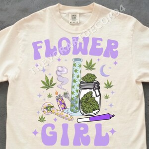 Könnte beinhalten: Cremefarbenes T-Shirt mit einem lila Aufdruck mit der Aufschrift "FLOWER GIRL". Das Design umfasst Cannabisblätter, ein Glas mit grünen Knospen, eine Pfeife, ein Feuerzeug, eine Bong und einen vorgedrehten Joint.