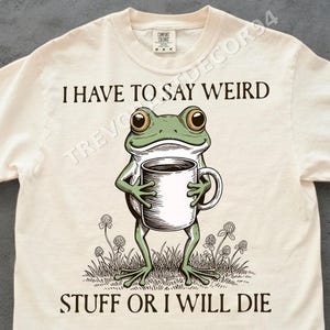 Könnte beinhalten: Cremefarbenes T-Shirt mit einer Grafik einer grünen Frosch, der eine Kaffeetasse hält. Der Frosch steht im Gras mit Klee. Der Text auf dem Shirt lautet: "I HAVE TO SAY WEIRD STUFF OR I WILL DIE."