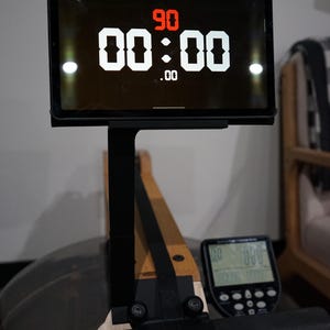 May include: A digital display on a rowing machine shows a timer with the numbers 90 and 00:00.00 in white against a black background. The display is mounted on a black stand, with a wooden rowing machine base.