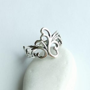 Könnte beinhalten: Ein silberfarbener Ring mit einem kunstvollen, wirbelnden Filigran-Design. Der Ring hat ein zentrales blütenähnliches Motiv mit zarten, geschwungenen Linien und offenen Räumen. Der Ring wird auf einer weißen, glatten Oberfläche präsentiert.