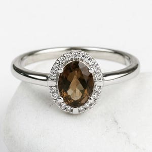 May include: A silver-toned ring featuring a central oval-cut smoky brown gemstone. The stone is surrounded by a halo of small, clear, round-cut stones set into the metal frame, which connects to a smooth, polished, solid silver-toned band.
