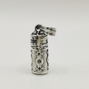 May include: A silver-colored, cylindrical pendant with intricate etched designs. The pendant has a hinged lid and a small ring for attachment. The body of the pendant features a repeating pattern of stylized shapes.