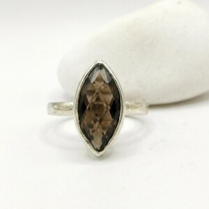 May include: A silver ring featuring a marquise-cut smoky quartz gemstone. The stone has a faceted surface with a geometric pattern and is set in a simple metal band. The ring is positioned upright, showcasing the elongated shape of the brown stone.