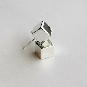 May include: A pair of silver stud earrings. Each earring has two stacked, geometric cubes. The top cube is slightly rotated, creating a dynamic visual effect. The earrings have a polished, reflective surface, and a post for pierced ears.