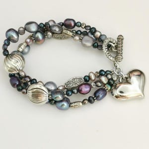 May include: A multi-strand bracelet with grey, purple, and silver beads. It features large, ribbed silver beads and a silver heart charm. The clasp is a decorative toggle.