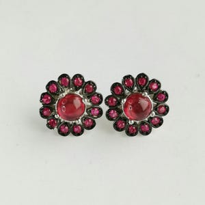 May include: A pair of stud earrings featuring a central round cabochon red stone surrounded by a halo of smaller faceted red stones set in a dark metal frame. The floral-shaped design highlights the contrast between the deep red gems and the dark setting.