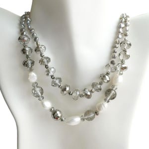 May include: A two-strand necklace displayed on a white mannequin. The necklace features clear faceted beads, gray beads, and white pearl-shaped beads. The necklace is silver in color and has a delicate design.
