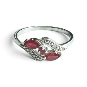 May include: A silver-toned ring featuring two marquise-cut red stones and one small round red stone in the centre. The band is accented with small clear stones along the curved metal setting that frames the red gemstones in a bypass design.