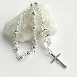 Op de afbeelding: Een zilveren rozenkransketting met een kruisvormige hanger. De ketting heeft kleine, ronde kralen die met fijne schakels zijn verbonden. Het kruis heeft een eenvoudig ontwerp en de ketting wordt getoond op een wit, getextureerd oppervlak.