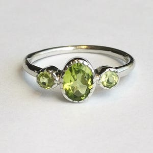 May include: A silver ring featuring a central oval-cut light green gemstone flanked by two smaller round-cut light green gemstones. The stones are set in a polished silver band with a scalloped metal edge surrounding each of the three green gems.