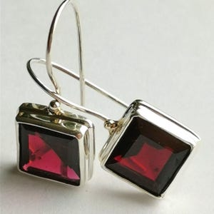 May include: A pair of silver drop earrings featuring square-cut, deep red garnet gemstones set in polished silver frames. The earrings are attached to curved silver ear wires, showcasing a clean, geometric design with a vibrant, dark red colour.