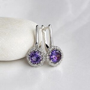May include: A pair of silver-toned drop earrings featuring a round, faceted purple gemstone at the centre of each. Each stone is surrounded by a halo of small, clear, sparkling stones set in a circular frame with a secure hinged lever-back closure.