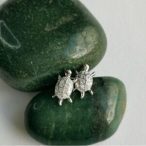 Könnte beinhalten: Ein Paar silberfarbene Schildkröten-Ohrstecker, besetzt mit kleinen, funkelnden Steinen. Die Ohrringe werden auf einem glatten, dunkelgrünen Stein präsentiert, was einen Kontrast in Texturen und Farben erzeugt. Das Schildkröten-Design verleiht einen verspielten Touch.