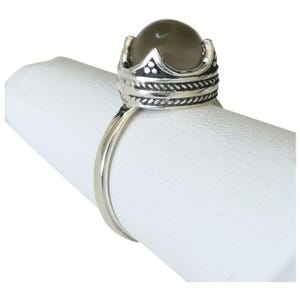 May include: A silver ring with a crown-shaped setting. The ring features a round, smoky quartz gemstone. The crown design has decorative details. The ring is displayed on a white surface.