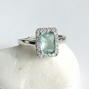 May include: A silver ring featuring a rectangular, light blue gemstone surrounded by small, clear stones. The ring is set on a white surface, showcasing the jewelry's design and sparkle. The ring's band is a simple, polished silver.
