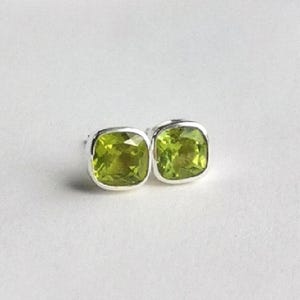 May include: A pair of square-cut peridot stud earrings. The earrings feature a bright, lime green gemstone set in a silver-coloured metal. The gemstones are cushion-cut and have a polished finish. The earrings are displayed on a white background.