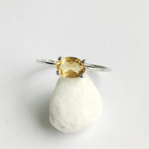 May include: A silver-toned ring featuring a central oval-cut yellow citrine gemstone held by four prongs. The band is thin and metallic, showcasing a polished finish. The ring is positioned resting on top of a smooth, rounded white stone.