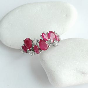 Puede incluir: Un anillo de plata con un grupo de rubíes de color rojo intenso en talla pera y marquesa, intercalados con pequeños diamantes blancos redondos y transparentes. Las piedras están engastadas en una banda metálica con diseño curvo.