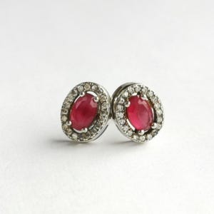 May include: A pair of ruby earrings. Each earring has an oval, deep red ruby stone, encircled by a halo of small diamonds. The earrings are set in a silver-coloured metal, offering a classic and elegant design. The earrings are suitable for everyday wear.