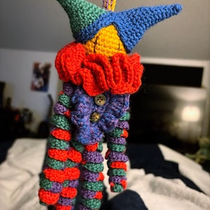 Könnte beinhalten: Eine gehäkelte Jesterpuppe mit einem mehrfarbigen Hut, einem roten Rüschenkragen und einem gestreiften Körper. Die Puppe ist aus grünem, blauem, gelbem, rotem und violettem Garn gefertigt.