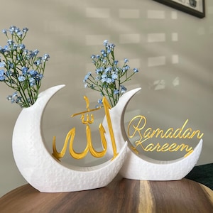 Könnte beinhalten: Zwei weiße, mondförmige Vasen aus Keramik mit goldener Kalligraphie. Die Vase links trägt das Wort "Allah" in arabischer Schrift. Die Vase rechts trägt den Text "Ramadan Kareem". Jede Vase hat einen Strauß hellblauer Blumen.
