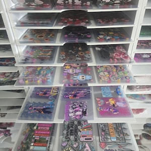 May include: A white storage unit filled with clear plastic sleeves containing various stickers. The stickers feature floral designs, cartoon characters, and text. The unit has multiple shelves and drawers, offering organized storage.