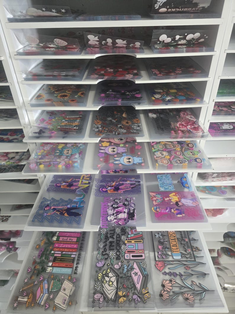 May include: A white storage unit filled with clear plastic sleeves containing various stickers. The stickers feature floral designs, cartoon characters, and text. The unit has multiple shelves and drawers, offering organized storage.